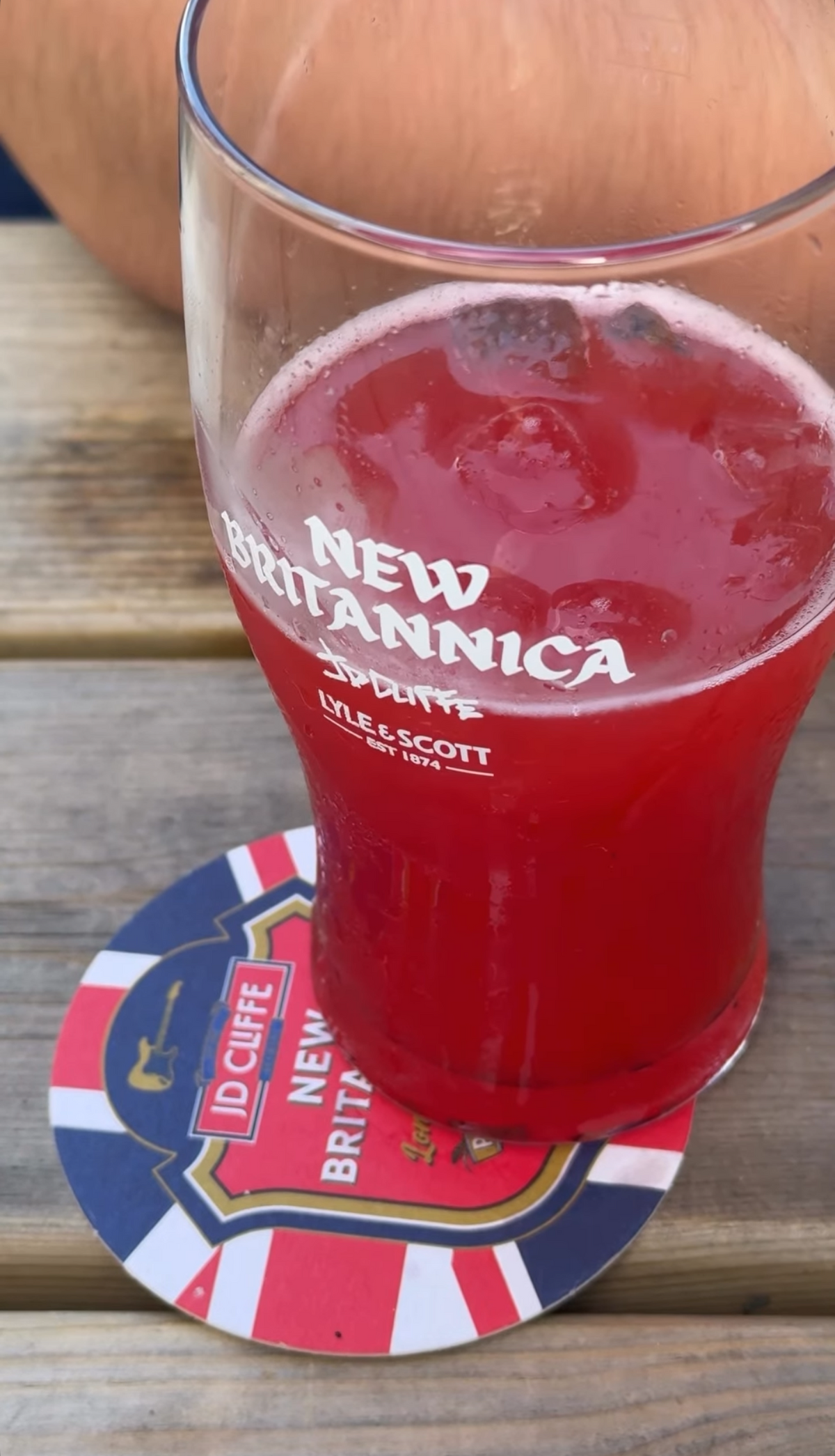 A pint glass filled with a red beverage, branded New Britannica, resting on a round New Britannica coaster on a wooden surface.