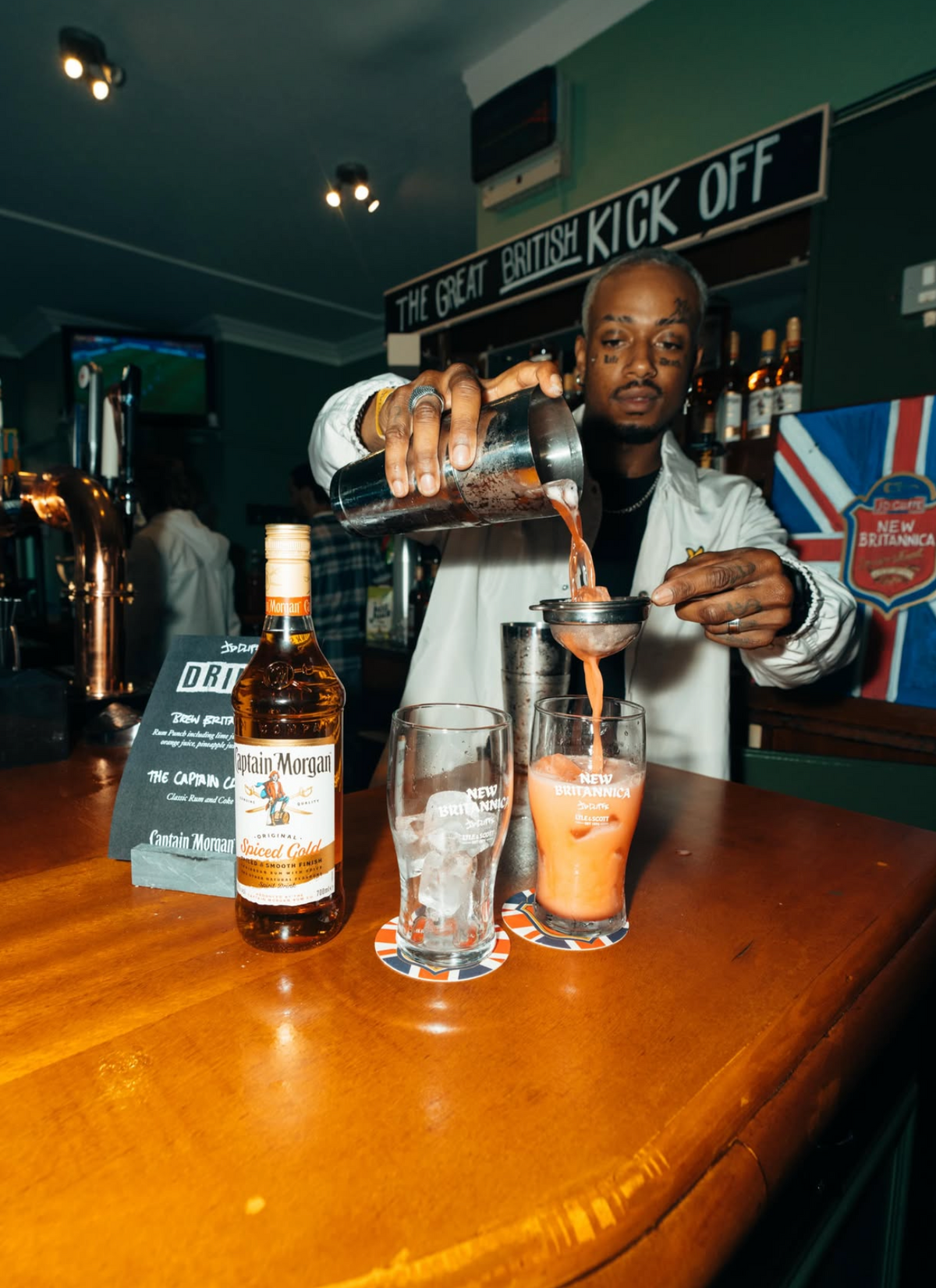 A bartender in a bar pouring a drink from a metal pitcher into two glasses on a wooden counter. A bottle labeled 'The Great British Kick Off' is visible on the bar, with a dark pub interior in the background.