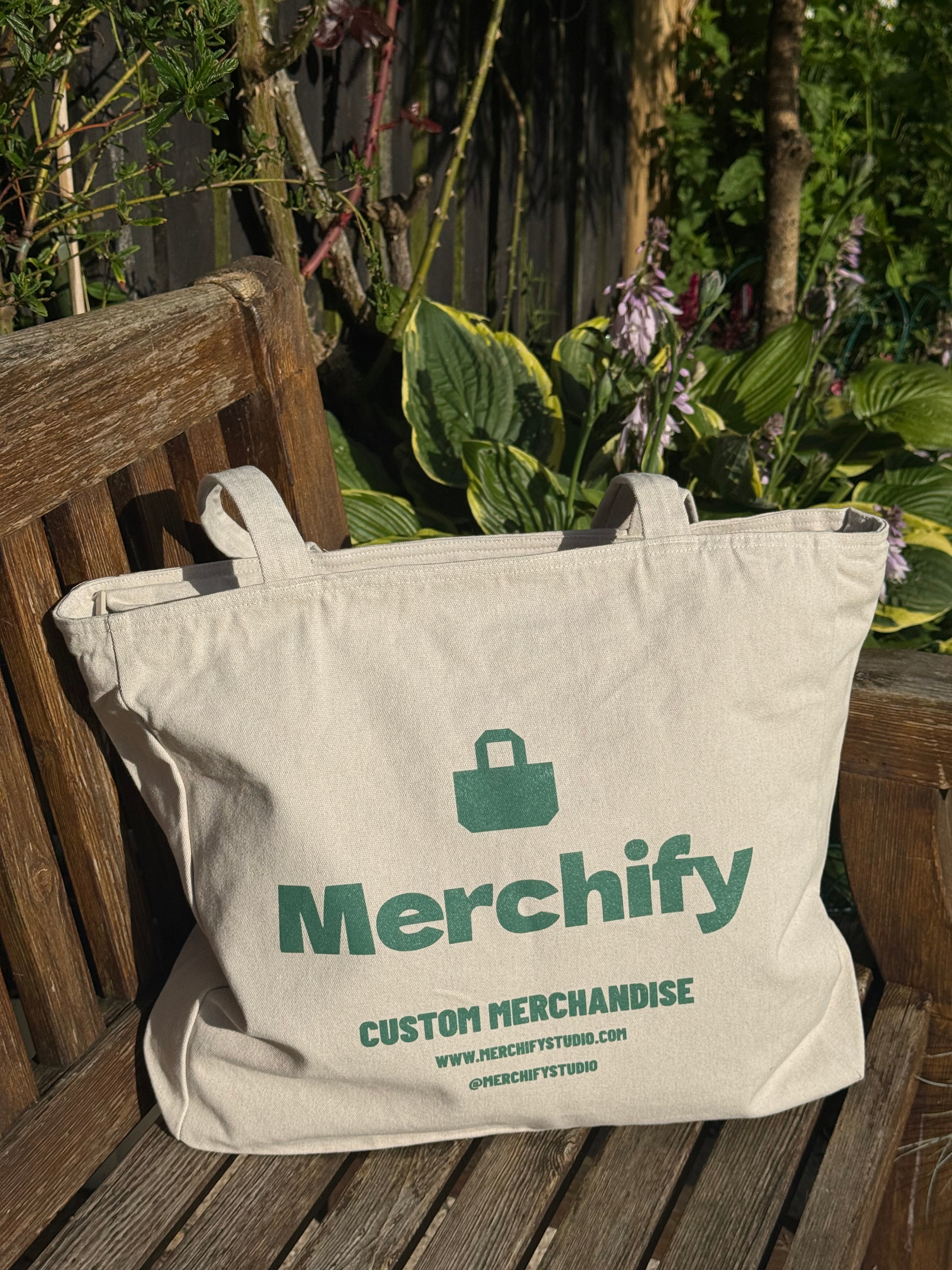 A natural canvas tote bag with green printed text reading 'Merchify' and 'Custom Merchandise' sits on a wooden garden bench with plants in the background.