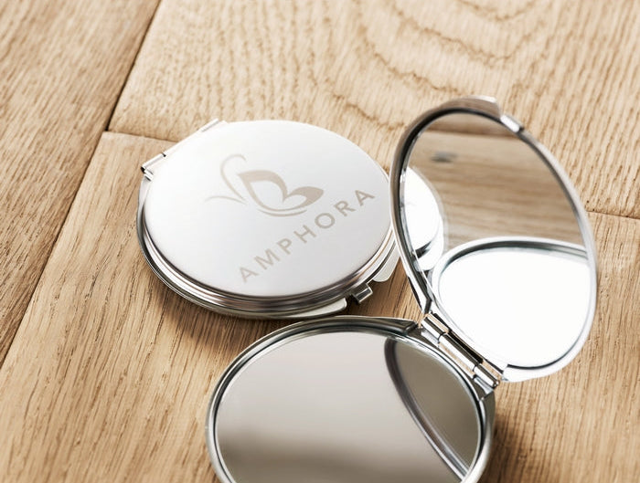 A silver compact mirror shown open and closed on a light wood surface. The closed mirror lid features the brand name 'AMPHORA' and a butterfly logo engraved on the front.