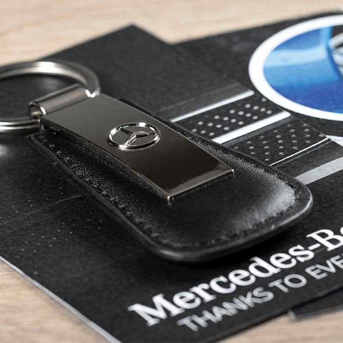 A black leather Mercedes-Benz key fob cover with a polished metal Mercedes-Benz star emblem, attached to a silver split ring, resting on branded black cardstock.