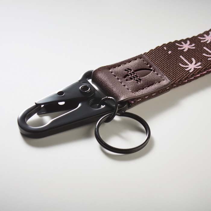 A close-up of a brown fabric strap with a black metal clasp and key ring, featuring a stitched patch with a stylized symbol and pink printed motifs on the fabric.