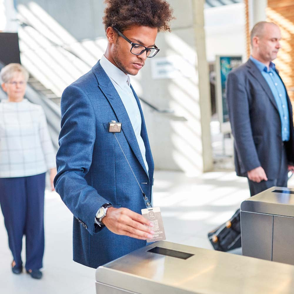 A man in a blue suit and glasses inserts a card into a metal turnstile at a building entrance, with another man and a woman standing in the background.