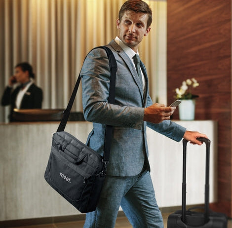A man in a gray suit walks through a hotel lobby while carrying a black shoulder bag with a white logo and pulling a rolling suitcase.