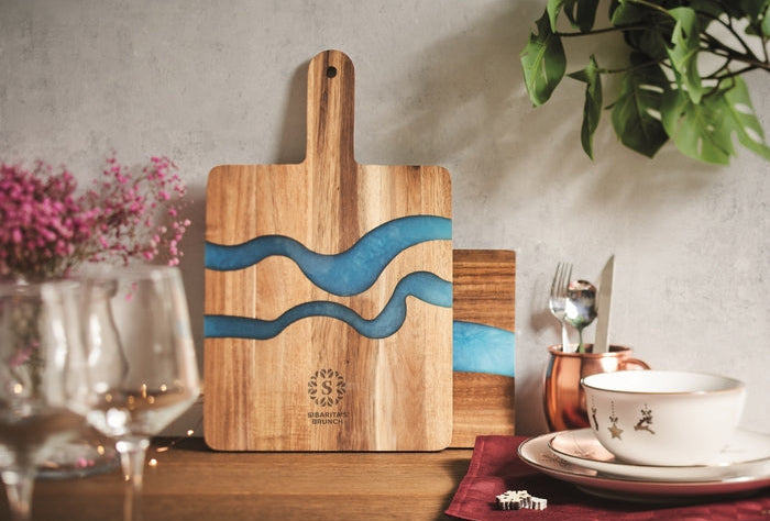 A wooden serving board with a handle and two wavy blue resin inlays, displayed on a tabletop in a styled kitchen setting with wine glasses, dishes, and a plant.