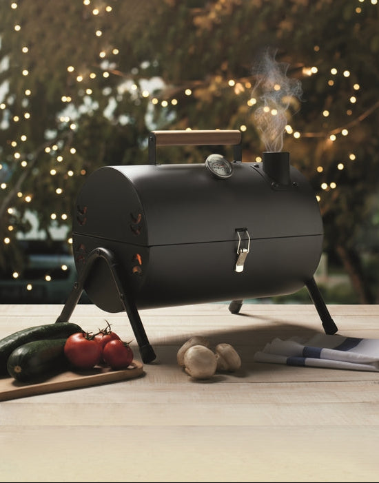 Black barrel-style barbecue grill with smoke, surrounded by vegetables on a wooden surface with a festive background.