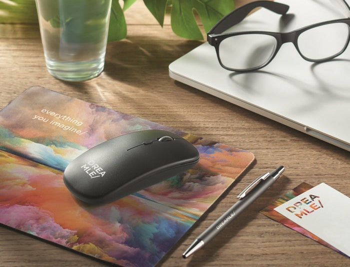 A black wireless computer mouse with 'DPEA MILE' branding resting on a colorful abstract mousepad on a wooden desk, alongside a silver pen, glasses on a laptop, and a glass of water.