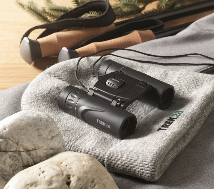Black Trek 28 binoculars resting on a gray knit pouch with the Trek 28 logo, surrounded by rocks and a pair of trekking poles on a wooden surface.