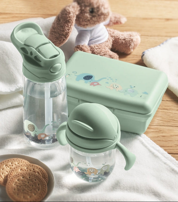 A set of baby feeding accessories including two small translucent bottles with pale green flip-top lids and a matching rectangular storage case, styled on a white cloth with a wooden surface, cookies, and a plush bunny in the background.