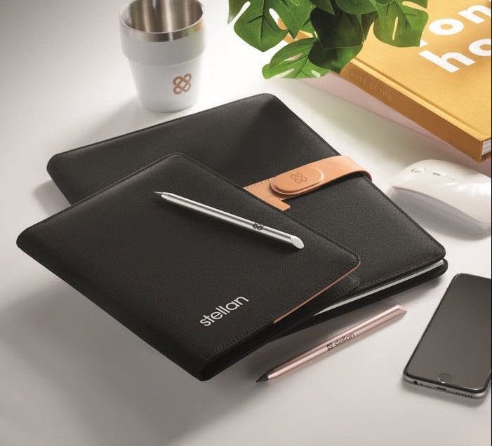 Two black folio-style tablet cases with tan leather accents are arranged on a light gray surface with office props, including a white cup, plant leaves, a book, a mouse, a smartphone, and pens. The front case shows the brand name 'stelian' in white near the bottom left.