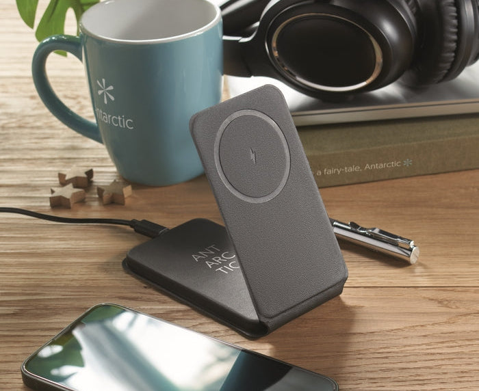 A dark gray ANT ANC wireless charging pad with a circular charging indicator, shown on a wooden desk next to a blue mug, headphones, and a smartphone.