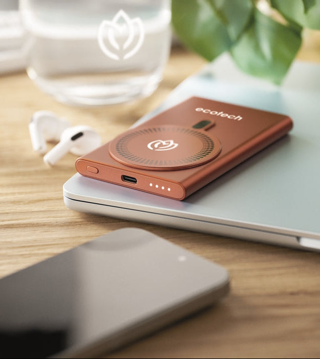 An Ecotech portable power bank in a metallic copper-orange finish, featuring a built-in wireless charger pad, resting on a silver laptop on a wooden desk with a glass of water, earbuds, and a smartphone nearby.