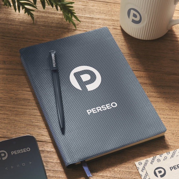 A dark blue textured PERSEO notebook on a wooden desk, styled with a matching PERSEO pen, a white PERSEO mug, and a smartphone.