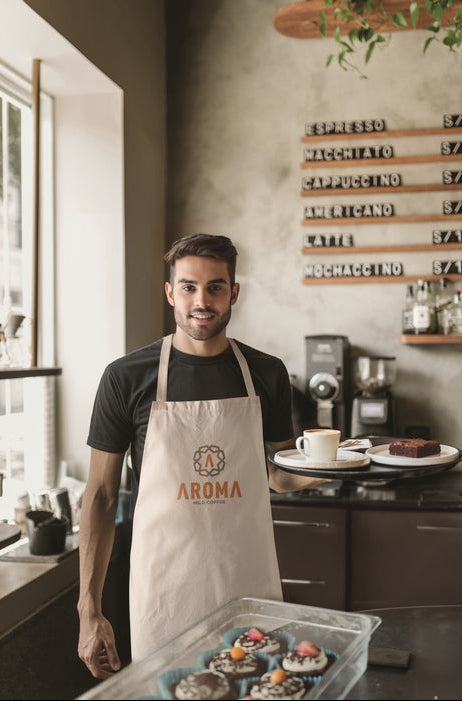 A man wearing a beige bakery apron with the brand name 'AROMA' stands in a kitchen setting, holding a tray of decorated cupcakes. The background features a rustic wall with wooden signs displaying coffee and tea options.