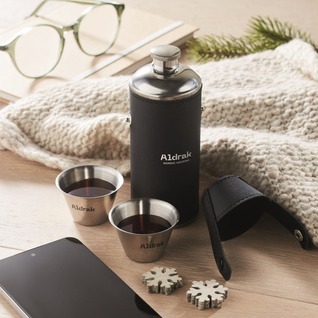 A black insulated flask with silver metal details stands on a wooden surface, surrounded by a cream knit blanket, green eyeglasses, a smartphone, two small metal cups, a black strap, and two snowflake-shaped accessories. The flask and cups are branded Aldrak.