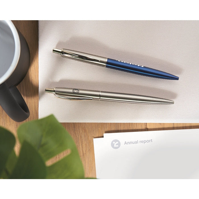 Two metal pens, one silver and one blue, placed on a white textured mat on a wooden desk with a plant leaf, a white report card, and part of a mug visible.