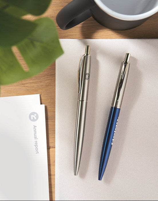 Two metal pens, one silver and one blue, placed on a white textured mat on a wooden desk with a plant leaf, a white report card, and part of a mug visible.