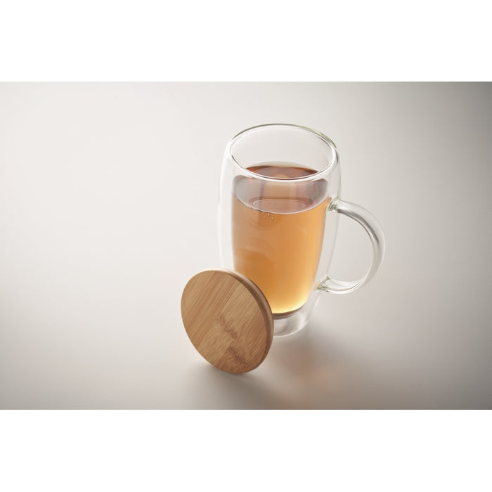 A clear double-walled glass mug filled with amber tea, shown with a round wooden lid leaning against the base on a light gray studio background.