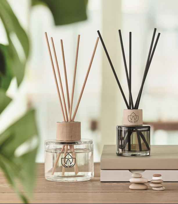 Two reed diffusers on a wooden surface, one with light natural reeds in a clear glass bottle and one with dark reeds in a dark glass bottle, both with light wood caps and a geometric logo, styled with books and small stones.