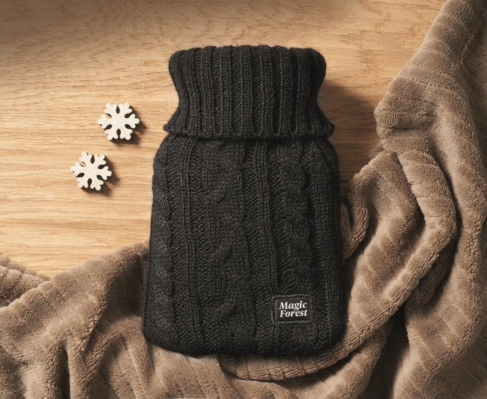 A black cable-knit hot water bottle with a ribbed neck, displayed on a brown textured blanket with two small white snowflake decorations on a wooden surface. A small label on the bottle reads 'Magic forest'.