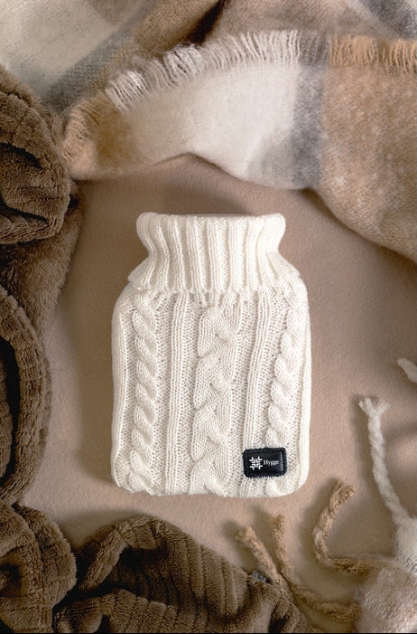 A cream-colored cable-knit hot water bottle cover with a small black brand tag, styled on a beige fabric background with brown and beige plaid blankets.