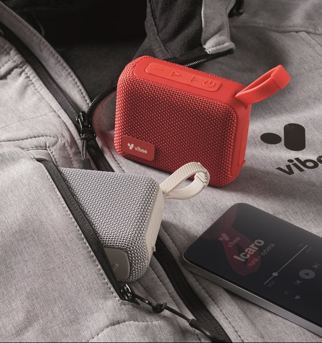 Two small Vibo portable Bluetooth speakers, one red and one gray, shown in a close-up lifestyle shot inside a gray zippered jacket pocket with a smartphone nearby.