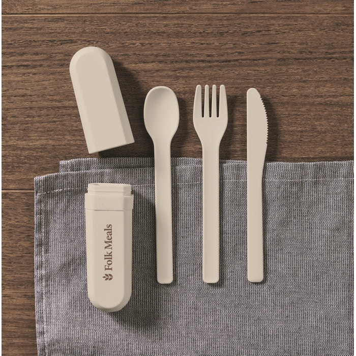 A 4-piece reusable cutlery set in a light beige color, including a spoon, fork, knife, and cap, displayed on a gray textured cloth over a dark wood background. The cap features the printed brand name 'Folk Meals'.