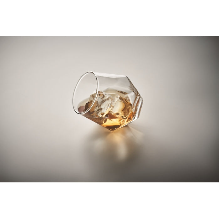 A clear faceted whiskey glass tipped on its side, containing an amber liquid and ice, set against a smooth gray gradient background.