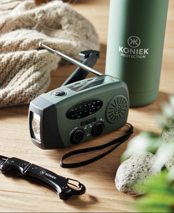A Konieck portable emergency radio in dark green and black sits on a wooden table with a matching green insulated bottle, a black wristlet strap, and a black folding knife nearby. A knitted blanket and stones are softly blurred in the background.