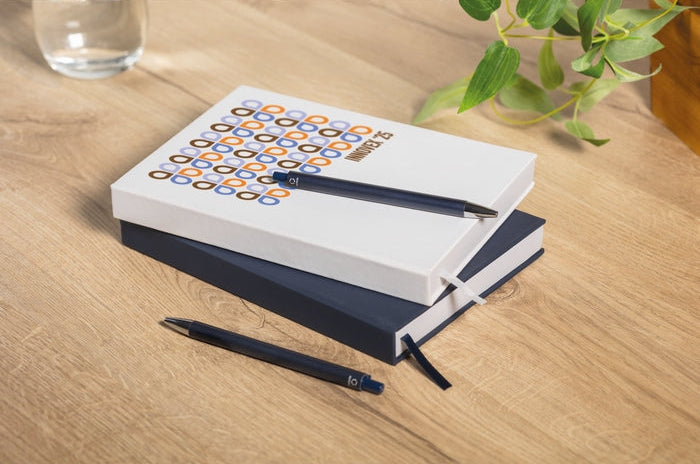 A stack of two notebooks on a light wood desk, with a white top notebook featuring a colorful geometric pattern and a dark navy bottom notebook. Two black pens are placed on top and beside the stack, with a glass and green plant leaves softly blurred in the background.