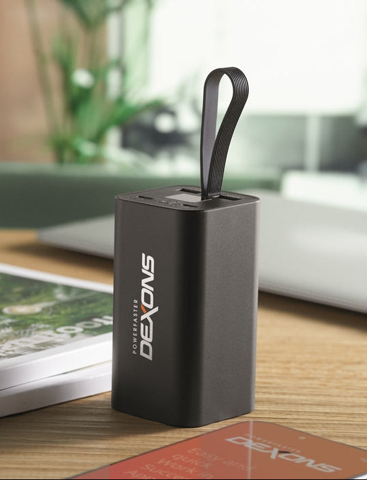 A black rectangular portable power bank with a loop handle, standing on a wooden desk. The front has the brand name 'Dexons' printed vertically in white.