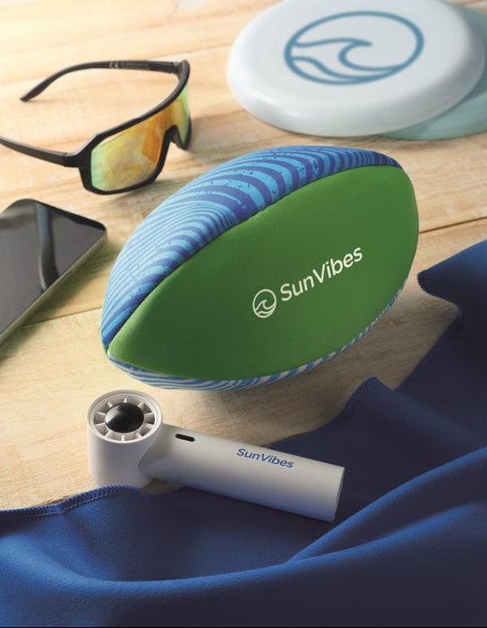 A SunVibes portable cooling fan and matching branded case are arranged on a light wood surface with a blue towel, sunglasses, a phone, and a frisbee nearby.