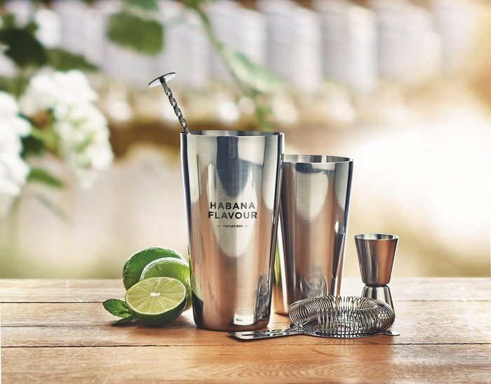 A stainless steel cocktail shaker set on a wooden table, featuring a tall shaker cup with 'HABANA FLAVOUR' printed on it, a smaller mixing tumbler, a jigger, a strainer, and lime slices, with a blurred outdoor patio background.