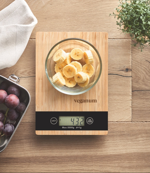 A wooden digital kitchen scale with a black display panel sits on a light wood tabletop. A clear glass bowl filled with sliced bananas rests on the scale, which shows 432 g.