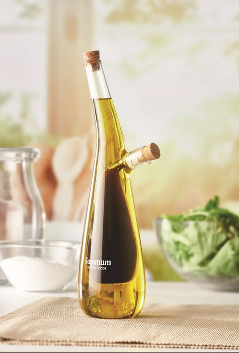 A clear glass bottle of olive oil with a cork stopper and a side branch spout, filled with golden olive oil. The bottle is set on a beige cloth with a blurred kitchen background showing a glass jar, white bowl, and a bowl of green salad.