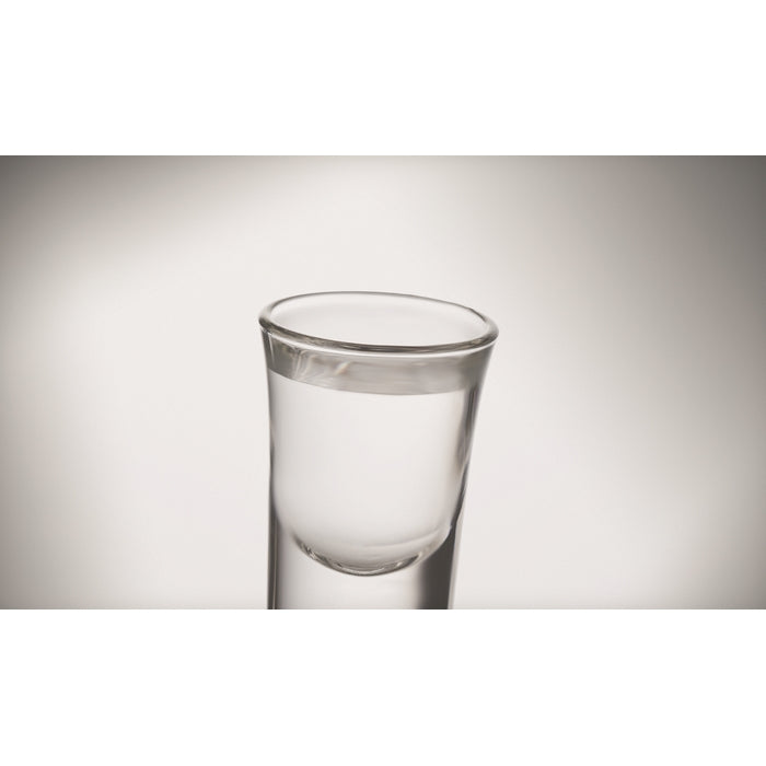 A clear glass shot glass centered against a neutral gray gradient background.