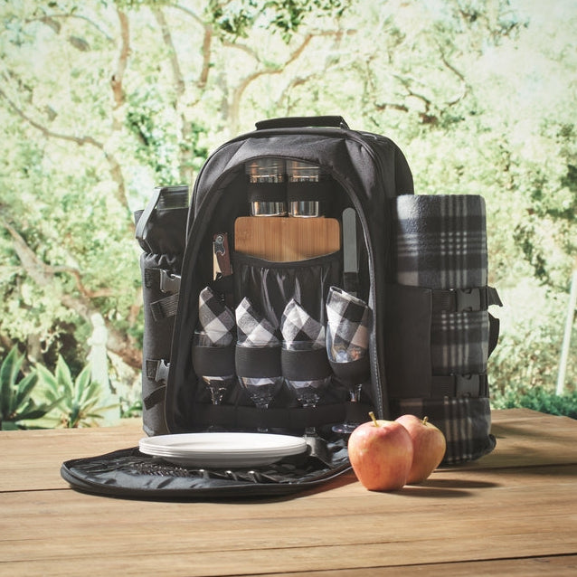 A black picnic backpack shown open on a wooden table, filled with a plaid blanket, glassware, bottles, and utensils, with two apples in front. Trees and a blurred outdoor setting are visible in the background.