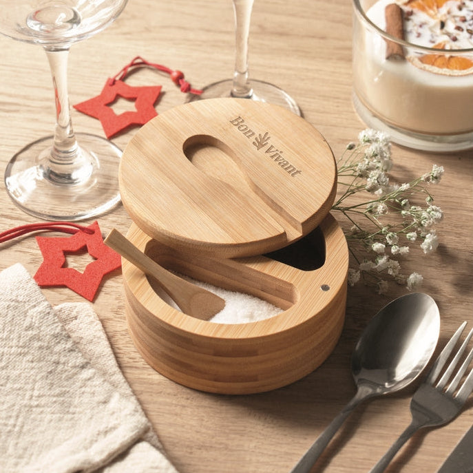 A round bamboo salt and pepper container with its lid off, showing a wooden spoon and white salt inside. The product is styled on a wooden table with wine glasses, a dessert, silverware, and small red star decorations.