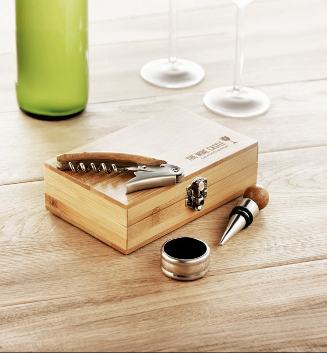 A wooden wine bottle opener set with a matching presentation box, shown on a light wood tabletop with a green bottle and wine glasses blurred in the background.
