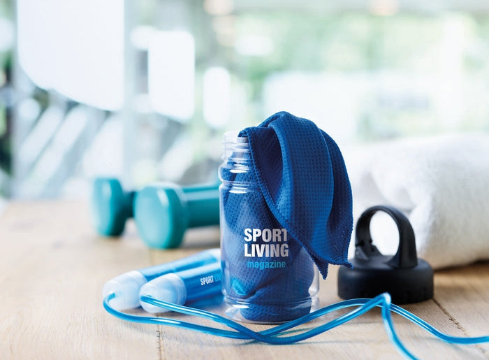 A blue textured towel draped over a clear water bottle with the text 'SPORT LIVING magazine' printed on it, placed on a wooden surface with dumbbells, a jump rope, and a towel in a softly blurred gym setting.