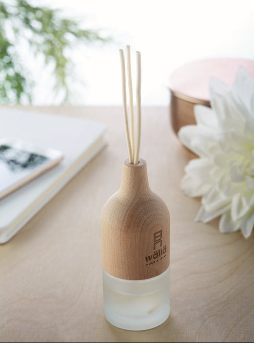A wooden reed diffuser bottle with a frosted glass base and three natural reeds, placed on a light wood tabletop with a blurred notebook, flower, and container in the background.