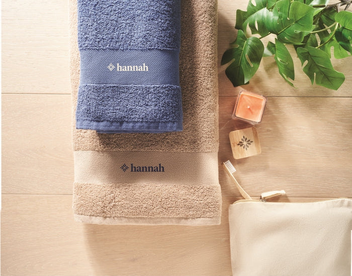 A styled flat-lay of two folded towels on a light wood surface: a blue towel on top with white 'hannah' embroidery, and a beige towel underneath with dark navy 'hannah' embroidery. A cream pouch, candles, and green leaves are arranged beside them as props.