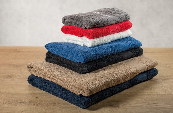 A stack of six folded cotton towels in various colors, including gray, red, white, blue, black, tan, and dark navy, arranged on a light wooden surface against a neutral gray wall.