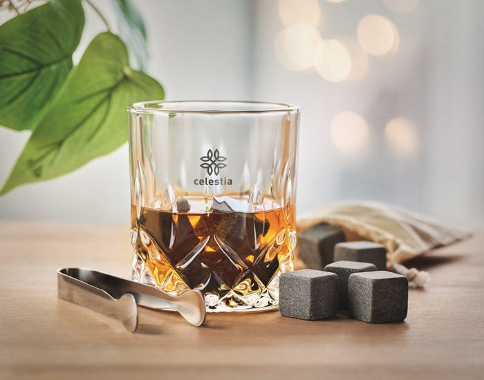 A clear cut-glass tumbler with the Celestia logo, partially filled with amber liquor, set on a wooden surface with ice cubes, metal tongs, and a small fabric bag in a softly lit indoor scene.