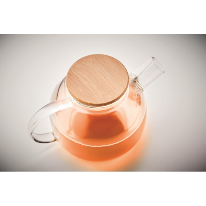 A clear glass teapot with a light wood lid, filled with an orange-amber liquid, shown on a neutral light gray background.