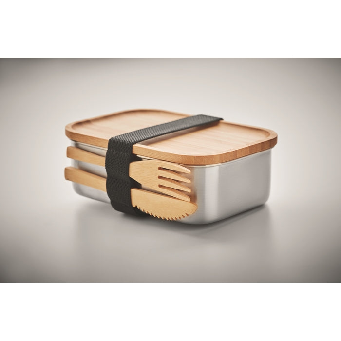 A rectangular stainless steel lunch box with a light wood lid, secured by a black elastic band holding two matching wooden utensils, shown on a neutral gray background.