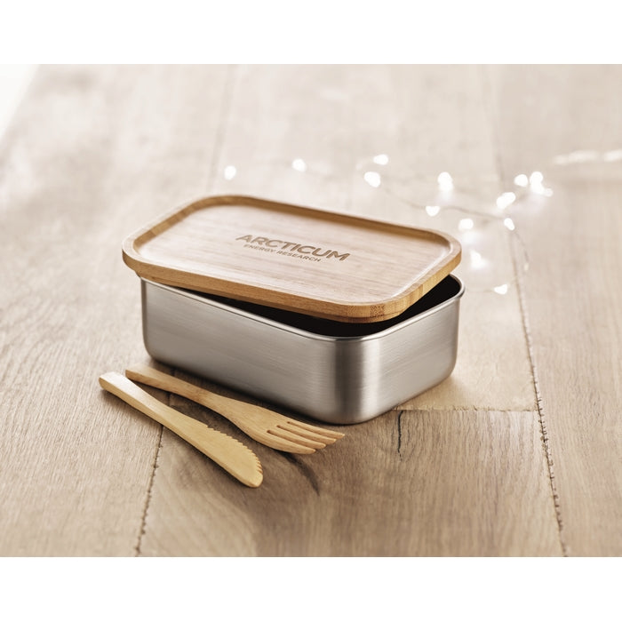 A rectangular metal lunch box with a bamboo lid, shown on a light wood surface with two matching bamboo utensils. The lid is printed with the brand name 'ARCTICUM'.