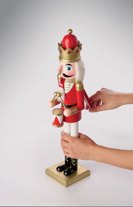 A red and gold toy nutcracker soldier on a gold base, with two hands adjusting its arm and holding its leg, against a light gray studio background.