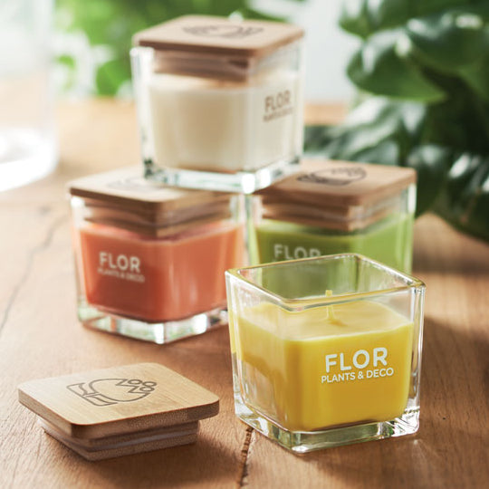 Set of square glass candles with wooden lids on a wooden surface with plants in the background