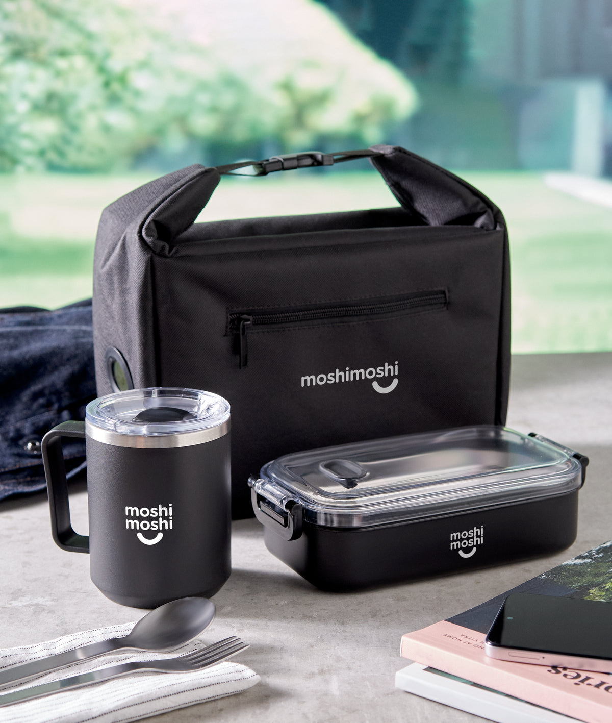 Black 'moshimoshi' lunch bag, mug, and food container on a table with a blurred green outdoor background.